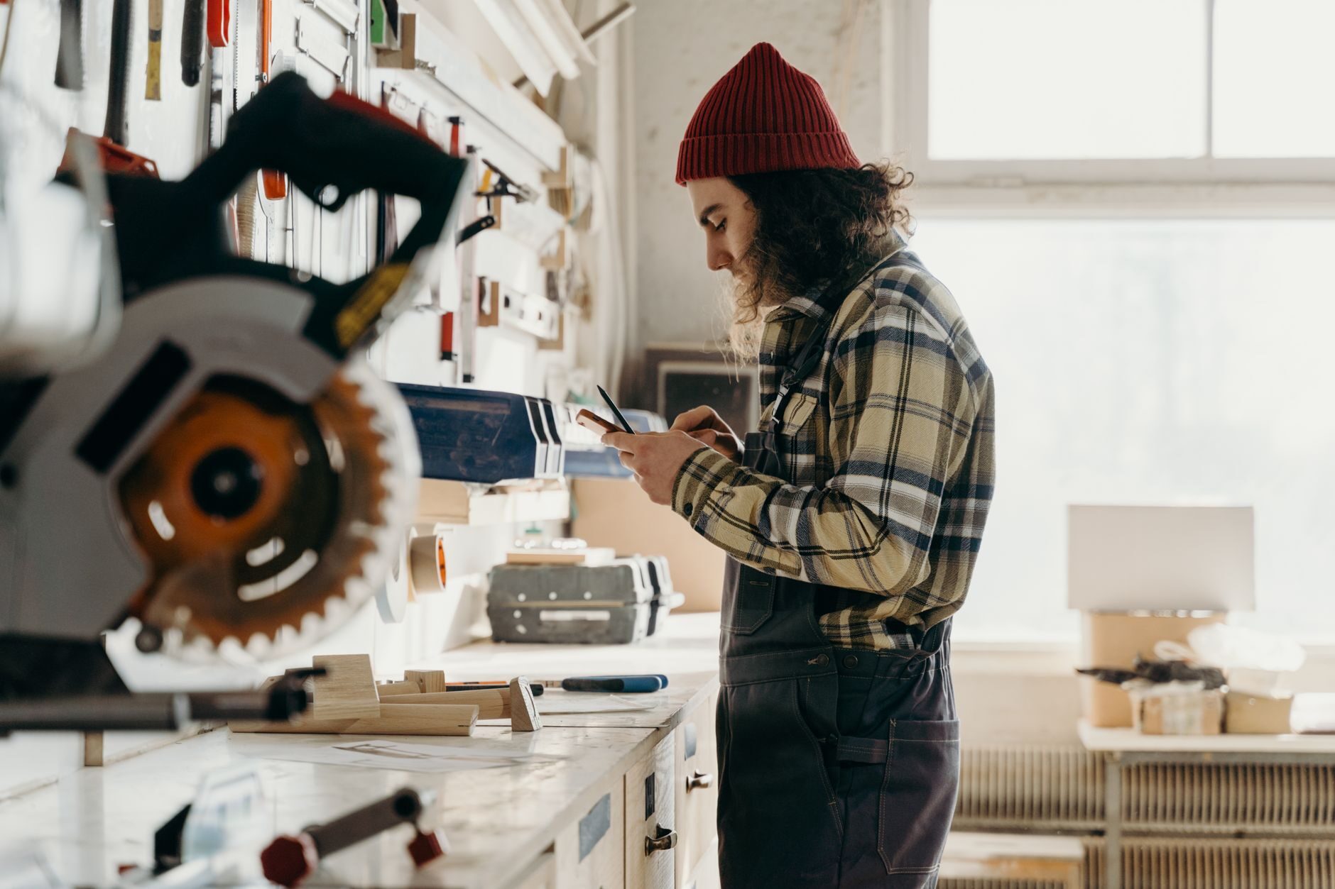 A tradesperson in flannel checking a smartphone in a woodworking workshop with tools in the background